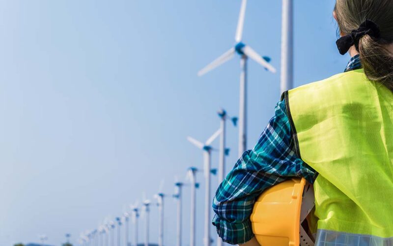 Woman engineer in uniform and holding yellow safety helmet with standing and checking wind turbine power